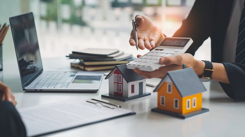 Person using calculator with small house models and laptop on table